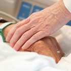 Photo of a person's hand touching an elderly patient's hand.